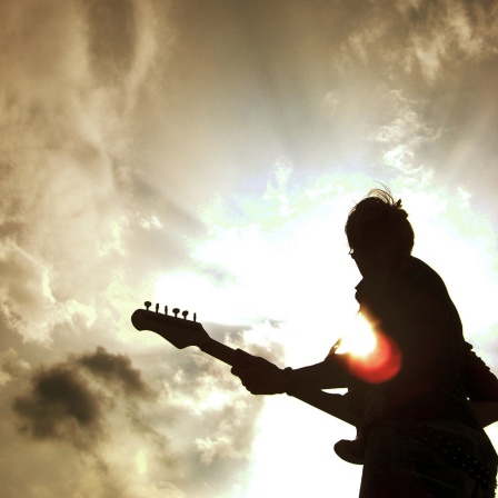 Die Silhouette einer Person mit Gitarre steht vor strahlend hellem Himmel. 