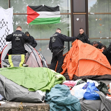 Polizisten räumen Zelte eines Protestcamps beiseite. An einer Wand hängt eine Palästina-Fahne.