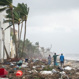 Nach einem Sturm ist ein Strand in Florida total verwüstet. Überall liegt Müll und Menschen gehen am Strand entlang.