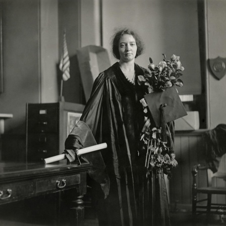 Die Physikerin Irene Joliot-Curie in akademischen Insignien: Sie trägt einen festlichen Talar, hält einen großen Blumenstrauß in der linken Hand und eine Schriftrolle in der rechten.
