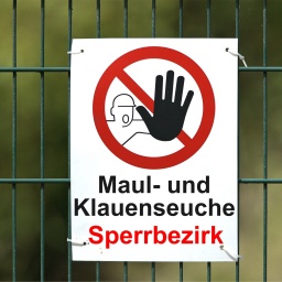 Ein weisses Warnschild mit der Aufschrift "Maul- und Klauenseuche Sperrbezirk" sowie einem Piktogramm mit einer Person mit ausgestreckter Hand in einem durchgestrichen roten Kreis an einem Doppelstabmattenzaun.