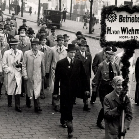 Eine Gruppe von Männern in Mäntel läuft zusammen eine Straße entlang. Der Gruppe voran gestellt läuft ein Junge, der ein Schild mit Hakenkreuz trägt auf dem "Betriebszelle H. von Wichmann" zu lesen ist.
