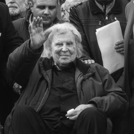 Mikis Theodorakis im Rollstuhl winkt in die Kamera.