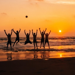 Eine Gruppe von Menschen springt am Strand vor einem Sonnenuntergang in die Luft sodass sich die Sihouetten der Personen klar abzeichnen.