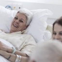 Symbolfoto: Eine weibliche Patientin im Krankenhausbett, rechts Besucher am Bett, links ein Arzt. Die Patientin lacht.