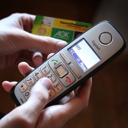 Ein Mann hält eine Gesundheitskarte der Allgemeinen Ortskrankenkasse (AOK) in der Hand, während er am Telefon eine Nummer wählt.