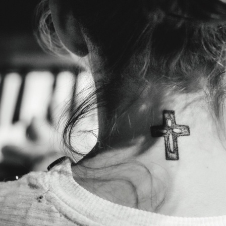 Eine junge Frau mit hochgesteckten Haaren sitzt am Klavier und spielt: Gezeigt wird sie in der Rückenansicht, so dass an ihrem Hals das Tattoo eines christlichen Kreuzes sichtbar wird. 