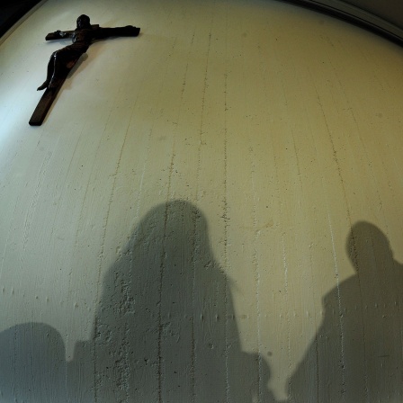 Schatten von Menschen an einer Wand, ebenso ein hängendes Kreuz