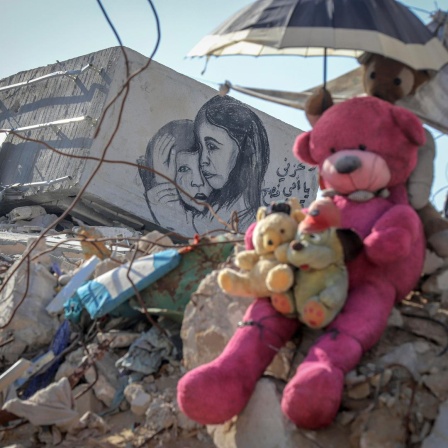 Teddybären sitzend auf einem Geröllhaufen eines eingestürzten Hauses in Gaza. Ein weiteres Stofftier hält einen kleinen Sonnenschirm über ihre Köpfe. Auf einem Stück Mauer ist eine Zeichnung auf der ein kleines Mädchen einen kleinen Jungen tröstend umarmt.