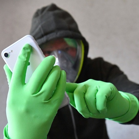Ein Person hält ihr Smartphone mit Schutzhandschuhen in den Händen und trägt eine Vollgesichtsmaske