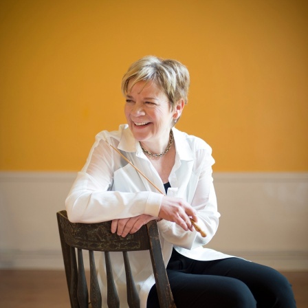 Porträt von Marin Alsop, seitlich auf einem Stuhl sitzend, zur Seite blickend mit freundlichem Lachen, vor gelbem Hintergrund.