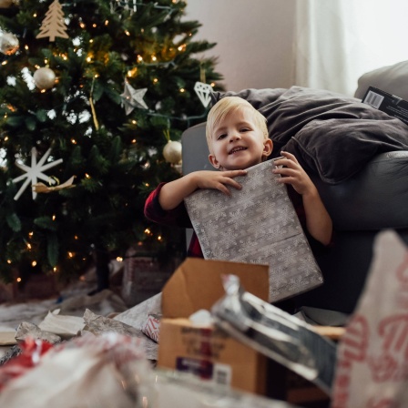 Kleiner Junge hält Geschenk hoch und lächelt vor dem Weihnachtsbaum