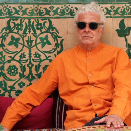 André Heller in orangefarbenem Hemd sitzt an einem Mosaiktisch