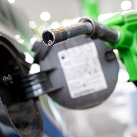 Eine Frau hält an einer Tankstelle an einer Zapfsäule eine Zapfpistole in der Hand und betankt ein Auto.