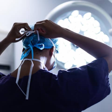 Eine Chirurgin ist im Operationssaal eines Krankenhauses von hinten zu sehen, während sie sich eine OP-Maske zubindet.