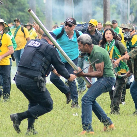 Ein Mann schlägt einen Polizisten mit einer Stange. Dahinter stürmen viele weitere Bolsonaro-Anhänger heran. 