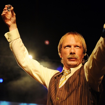  Der Musiker Achim Hagemann als Pawel Popolski auf der Bühne im Scheinwerferlicht, er hält ein Wodkaglas und in der anderen Hand ein Mikrofon.