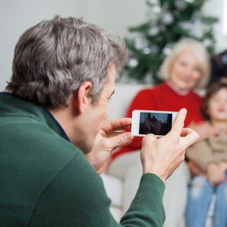 Ein Mann fotografiert mit einem Smartphone seine Familie vor dem Weihnachtsbaum. 