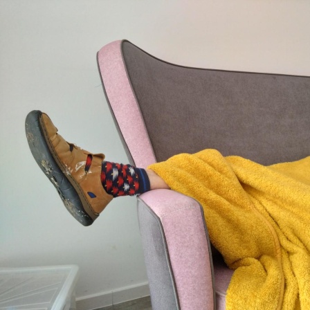 Eine Person liegt zugedeckt auf einem Sofa. Unter der Decke schaut ein Fuß heraus, der in einem Hausschuh und bunt gemusterten Socken steckt.