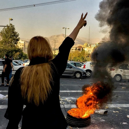 Proteste im Iran: Von hinten ist eine Frau zu sehen, sie trägt kein Kopftuch und reckt ihren Arm in die Höhe: Neben ihr brennt ein Feuer. 