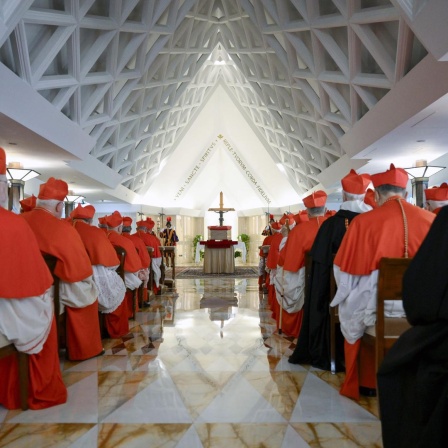 Bischöfe sitzen auf Stühlen in einer Halle mit spitzen Dach. An deren Ende steht ein Altar mit Jesus-Kreuz. 