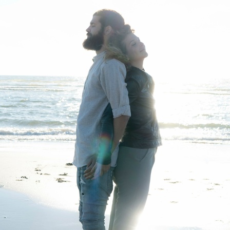 Paar steht mit geschlossenen Augen und Rücken an Rücken am Strand