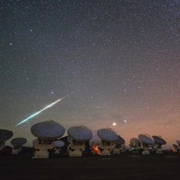 Meteorschweif am Sternenhimmel vor der Silhouette einer Teleskopanlage mit großer Radarschüssel.
