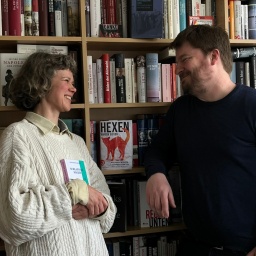 Lisa Kreißler und Alexander Solloch stehen sich zugewand an einer Bücherwand gegenüber. 