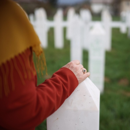 Die Hand einer Frau, die einen Ring trägt, liegt auf einem weißen Kreuz. Im Hintergrund sind weitere weiße Kreuze, die Gräber markieren zu sehen.