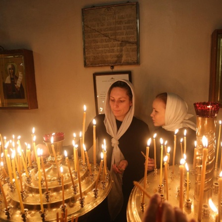 Zwei Frauen mit je einem Tuch über dem Kopf zünden während eines orthodoxen Gottesdienstes in der Kazaner Kathedrale in St. Petersburg Kerzen an, hinter ihnen sind Ikonen zu sehen.