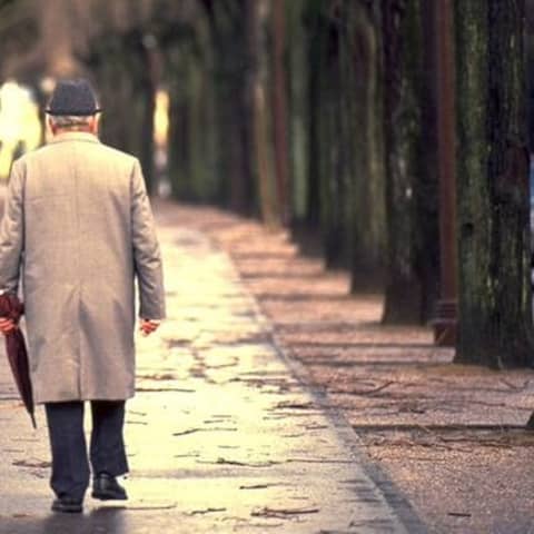 Ein älterer Herr mit einem Regenschirm läuft an einem Herbsttag alleine auf dem Gehweg.