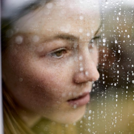 Eine junge Frau schaut traurig durch ein Fenster auf dem Regen sichtbar ist.