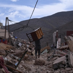Ein Mann trägt ein großes Möbelstück auf der Schulter. Um ihn herum Geröll, im Hintergrund das Atlasgebirge.