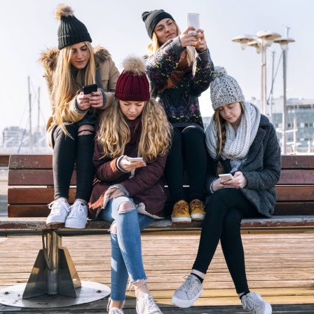 Vier junge Mädchen sitzen auf einer Bank und spielen mit ihrem Handys.