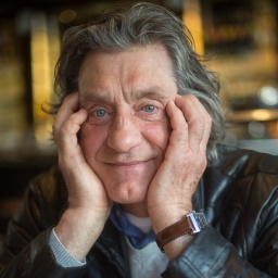 Winfried Glatzeder, ein alter Mann mit langen, grauen Haaren hat das Gesicht in die Hände gestützt und lächelt in die Kamera. 