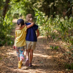 Zwei Kinder gehen Arm in Arm einen Waldweg entlang. 
