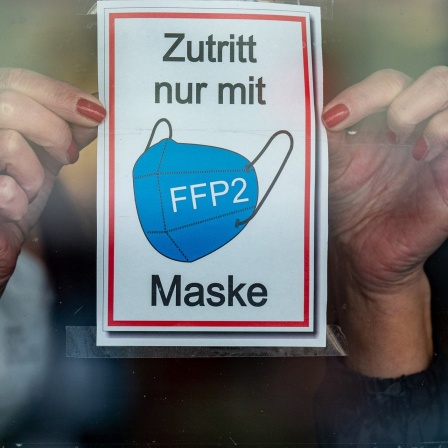 Eine Frau klebt ein Hinweisschild mit der Aufschrift „Zutritt nur mit FFP2 Maske“ an eine Glastür am Eingang eines Geschäfts 