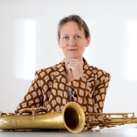 Ein Porträt einer an einem Tisch sitzenden Frau. Vor ihr auf dem Tisch liegt ein Tenorsaxofon.