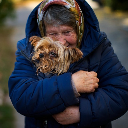 Die alte Frau in blauer Winterjacke und Kopftuch steht auf einer Straße und wärmt ihren kleinen Hund unter dem Mantel.