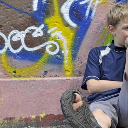 Lustloser neunjähriger Junge sitzt vor einer Graffiti-Wand, einen Fuß von sich gestreckt, sodass die kaputte Sohle des Schuhs sichtbar wird. (Symbolbild)