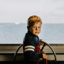 Ein Junge am Steuerrad eines Schiffs. Durch die Scheibe sieht man den Horizont und das Meer.