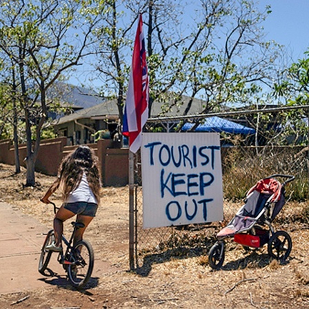 Eine Frau fährt Fahrrad auf Hawaii, neben ihr ein Protestschild mit dem Text: "Toursit keep out"  