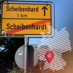Auf einem gelben Ortsschild steht durchgestrichen "Scheibenhardt" in deutscher Schreibweise mit "dt" und der Verweis auf den unmittelbar angrenzenden Ort Scheibenhard - ohne "t" geschrieben - in Frankreich, während im Hintergrund Fachwerkhäuser und Geranien zu erahnen sind.