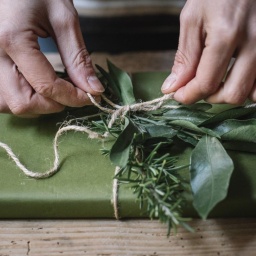 Die Aufnahme zeigt Frauenhände, die liebevoll ein in grünes Papier eingewickeltes Geschenk mit grünen Zweigen dekoriert.