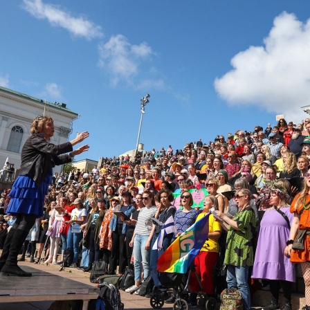 Eine Frau dirigiert eine Menschenmenge unter freiem Himmel, die auf einer großen Treppe steht und gemeinsam singt
