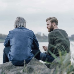 Zwei Menschen sitzen auf Steinen am Wasser und führen ein Gespräch