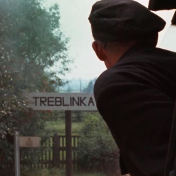 Szene aus Claude Lanzmanns Shoa. Eine Person guckt aus einem Zug, während dieser ein Schild mit der Aufschrift "Treblinka" passiert.