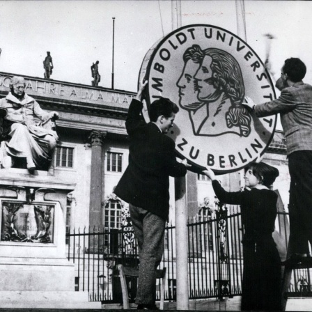 Drei Studierende halten vor der Humboldt-Uni zusammen ein großes, rundes Uni-Logo in der Hand.