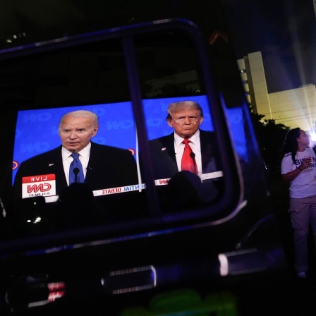 Bilder von US-Präsident Joe Biden und seinem republikanischen Konkurrenten Donald Trump spiegeln sich in einer offenen Autotür, während Zuschauer eine TV-Wahlkampfdebatte auf einer riesigen Leinwand in einem Freiluftkino verfolgen.