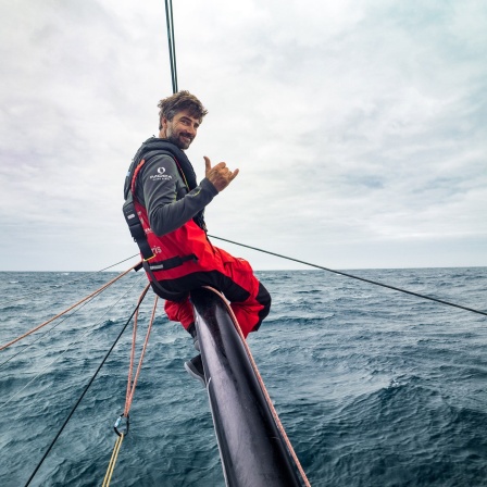 Boris Herrmann sitzt auf einer Stange am Bug der Malizia und lässt die Beine über dem Wasser baumeln. Er lächelt in die Kamera und zeigt Daumen und kleinen Finger zum hawaiianischen Gruß in die Kamera